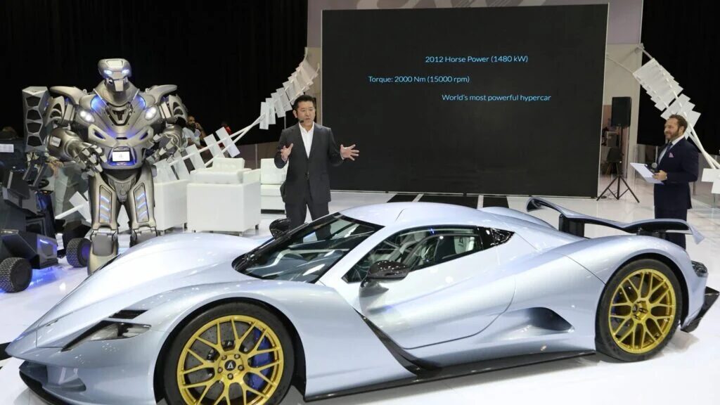 Titan the Robot stands with a corporate speaker on a trade stand, with the worlds fastest electric hyper car, the aspark owl.