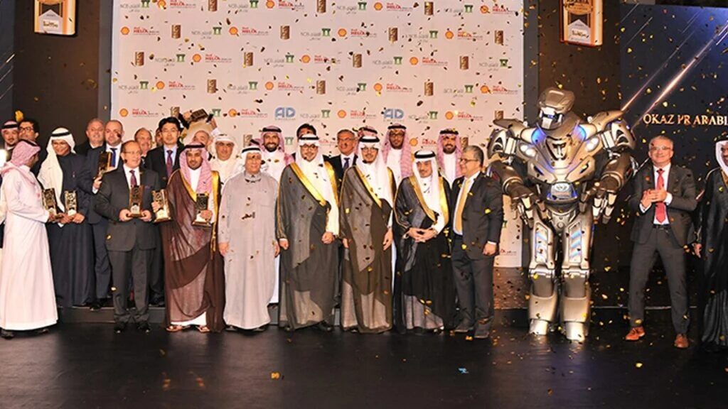 Titan the Robot stands on stage with a large team of arabic men. Gold confetti is coming from above and several of the men hold awards.