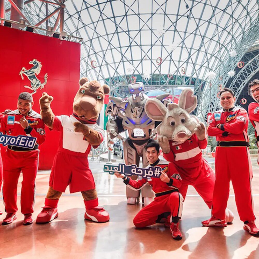 Titan the Robot stands with 2 mascots (one bunny and one camel) and 5 men. All are wearing red, motorsport all in ones. A ferrari logo can be seen on a banner and the staff hold Joy of Eid signs.