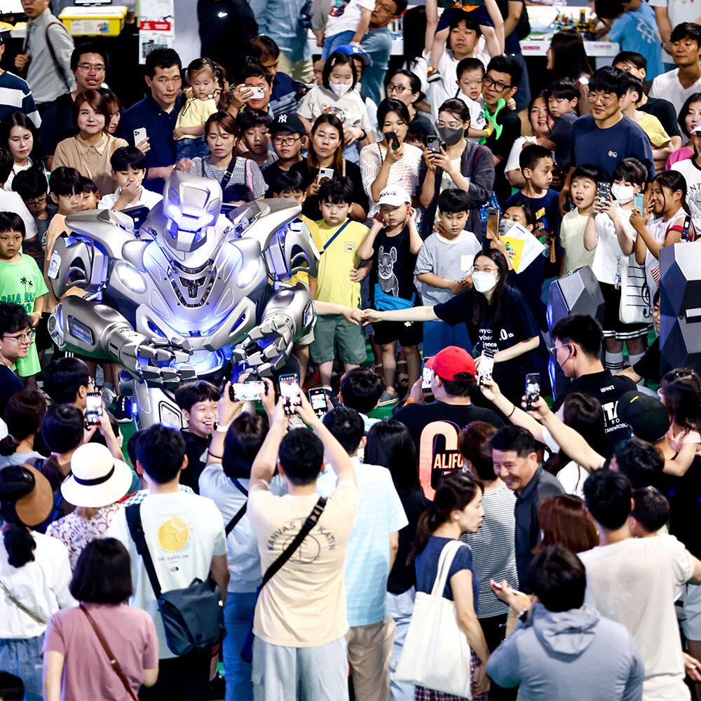 titan the robot is surrounded by a large group of school aged children at a trade show in south korea