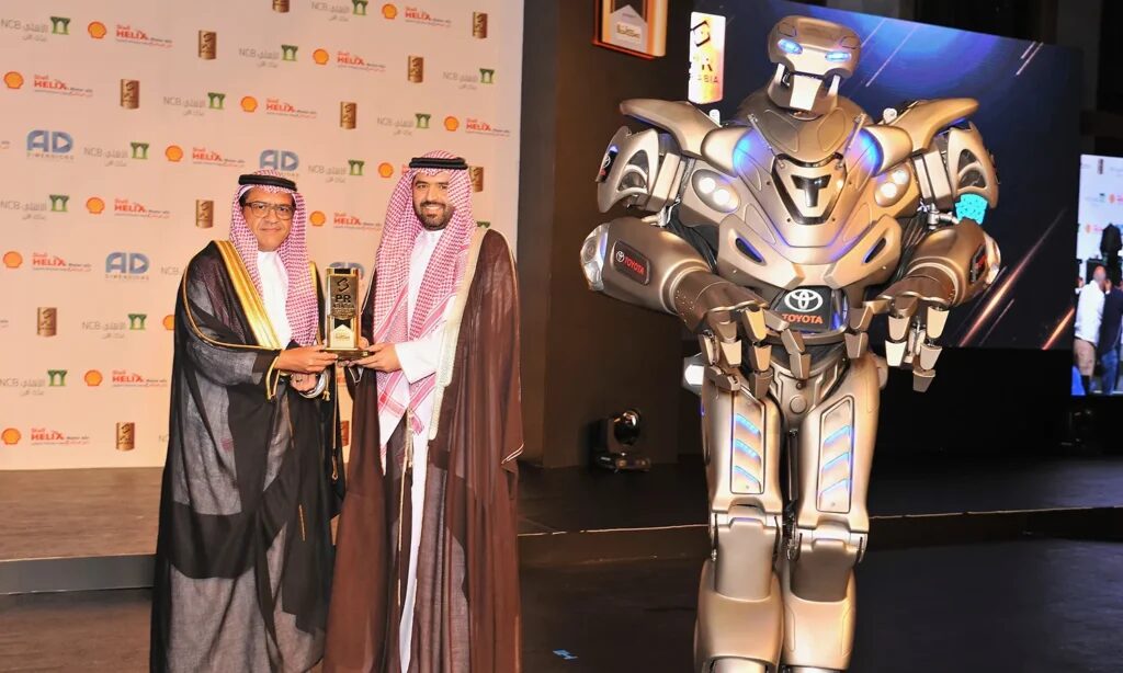 Titan the Robot stands on stage at an awards ceremony, with Toyota's logo on his chest. 2 men stand holding an award stood beside him.