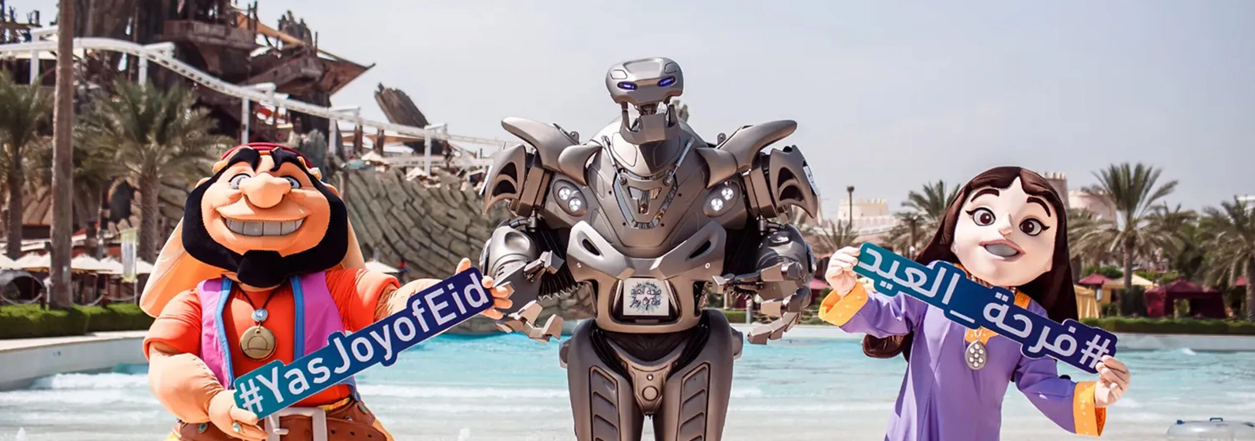 Titan the Robot poses for a photo with two mascot characters at the yas island water park. The characters are hold joy of eid signs.