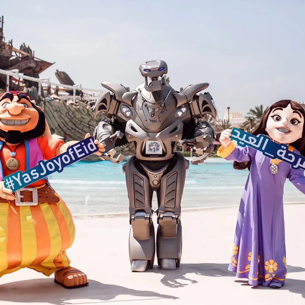 Titan the Robot poses for a photo with two mascot characters at the yas island water park. The characters are hold joy of eid signs.