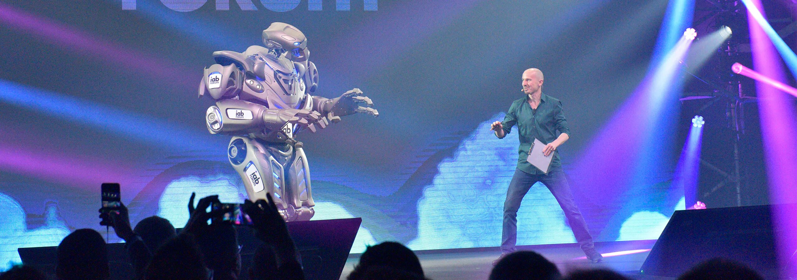 Titan the Robot stands on a stage, interacting with a presenter. He is branded with IAB Forum branding