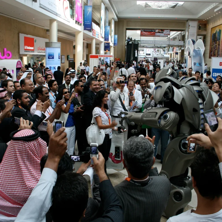 A large corporate crowd has gathered in the atrium of a large trade show. Nearly all have their cameras filming, while one woman stands close to titan and laughs.