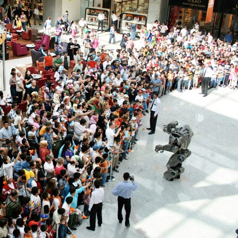 Titan the Robot performs to a large crowd in a shopping centre. The performance has shiny white tiled floor. shops are seen in the background