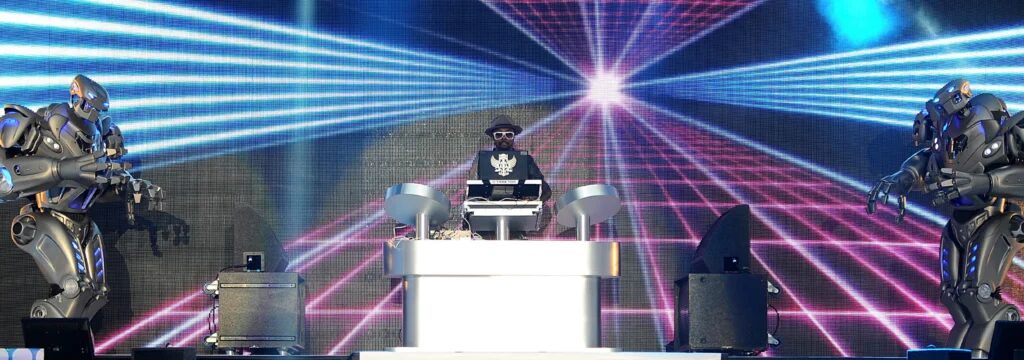 Will.i.am stands behind a dj desk, with a titan the robot on each side that look towards the camera.