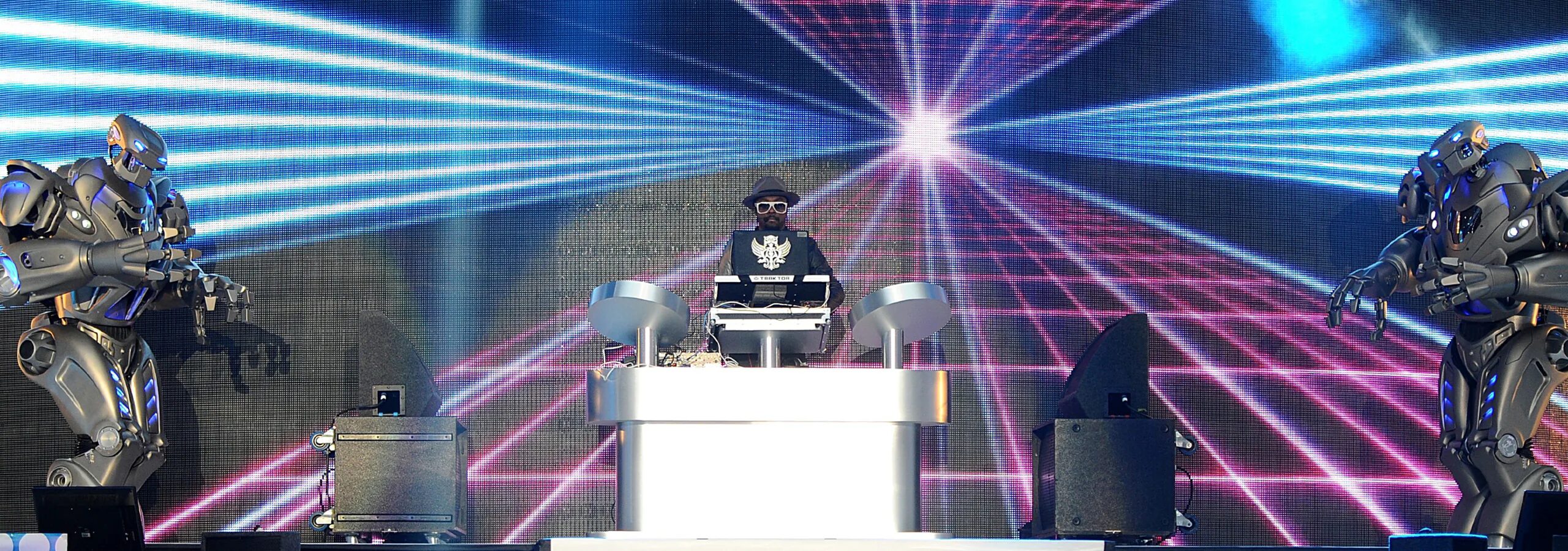 Will.i.am stands behind a dj desk, with a titan the robot on each side that look towards the camera.