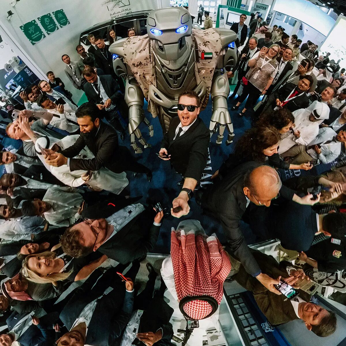 Titan the Robot and Dave take a selfie with a fish eye lens. A trade show crowd are gathered around and Titan is wearing a saudi arabian army jacket