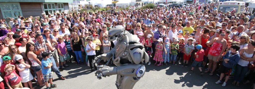 Titan the Robot performs to an outdoor crowd, that stands as far as the eye can see. An ice cream van can be seen amongst the crowd.