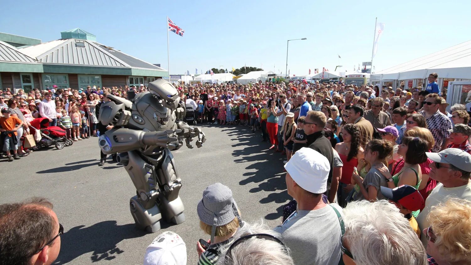 Titan the robot performs at an outdoor show to a huge crowd. the sun is shining and there is a union jack flag flying from a pole.
