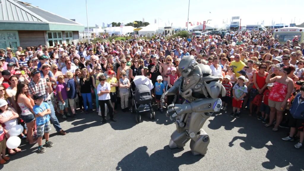 Titan the Robot performs to an outdoor crowd, that stands as far as the eye can see. An ice cream van can be seen amongst the crowd.