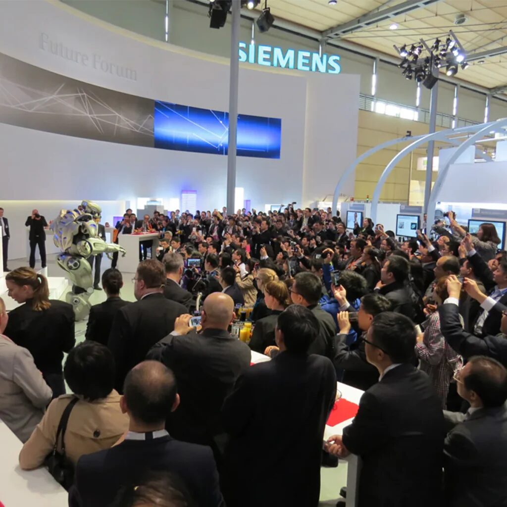 A large crowd has gathered around a Siemens trade show stand. Titan can be seen on the stand, with future forum on the backdrop.
