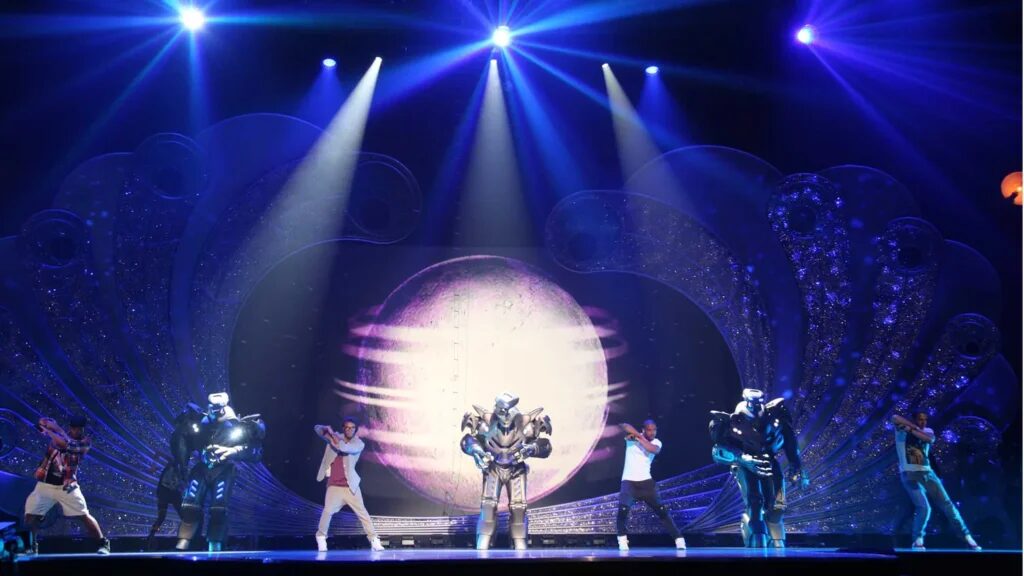3 Titan the Robots stand on stage with casually dressed members of JLS. A large image of a moon is projected onto the set, with blue and white spotlights from the ceiling.