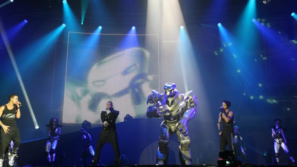 Titan the Robot stands between the boyband JLS as they sing on stage. A large image of titans face is seen on a screen behind, with several backing dancers.