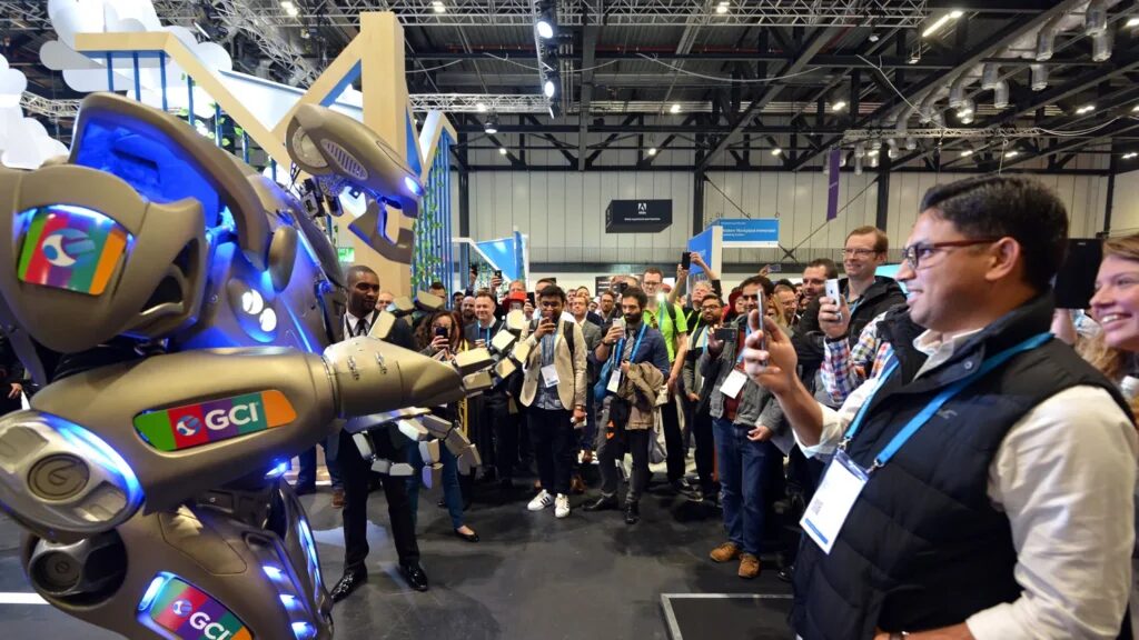 Titan the Robot performs to a trade show crowd who all have phones and are filming. Titan has white and blue, ARuVR branding on his shoulder and arm