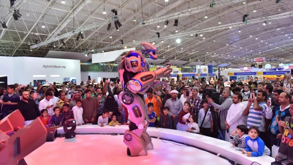 Titan the Robot stands on a small circular stage at a trade show. A large crowd have surrounded it. He has Toyota branding panels on the shoulder, arms and legs.