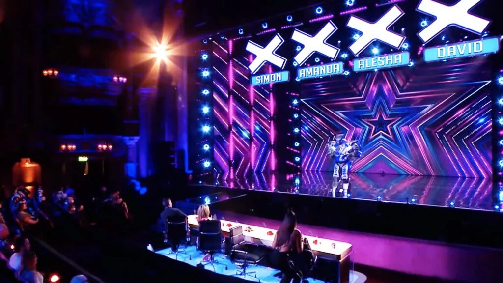 Titan the Robot stands on a stage underneath the famous X's of Britains Got Talent. 3 judges watch, with one empty chair.