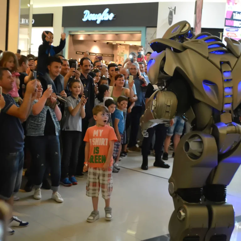 Titan the Robot stands in front of a crowd of people in a shopping mall corridor. One young boy in an orange t-shirt has stood closer to Titan and looks up in amazement.