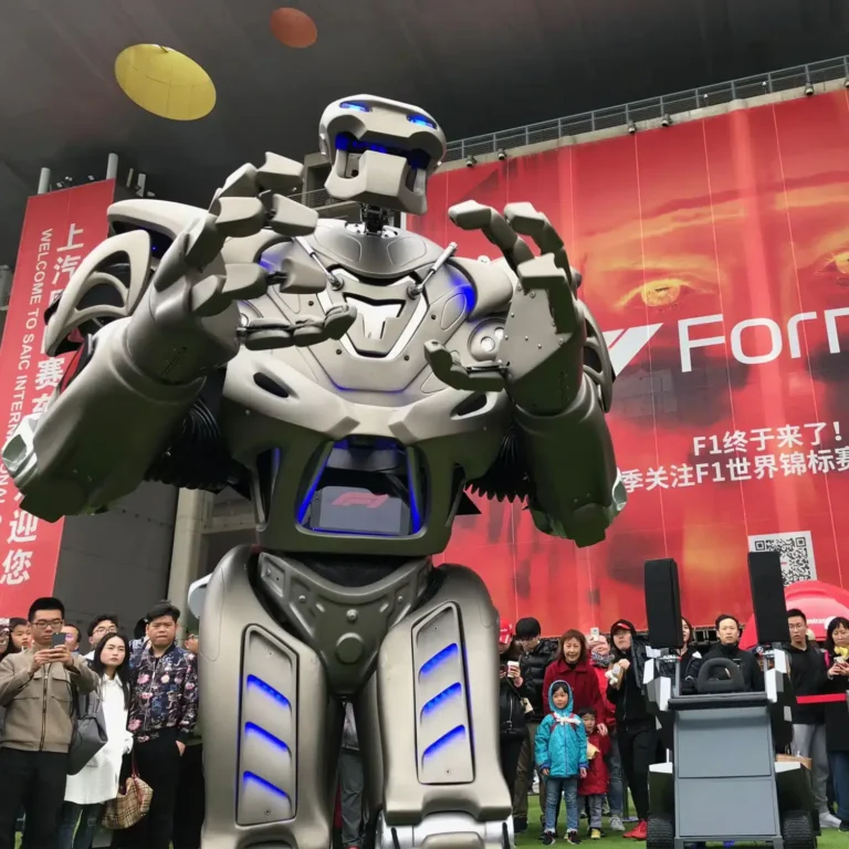 The camera looks up at Titan the Robot who looks out behind the camera. A large, red, formula one sign is behind a crowd, with chinese writing.