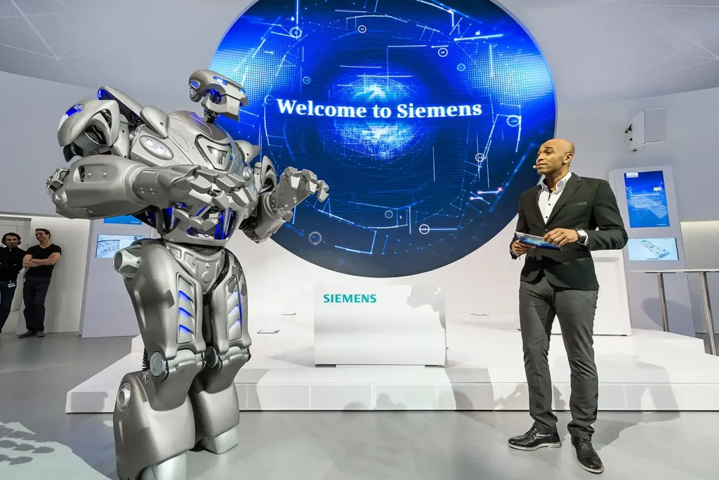 Titan the Robot and a presenter interacting with each other in front of a large, futuristic, digital display with the words "Welcome to Siemens" on.