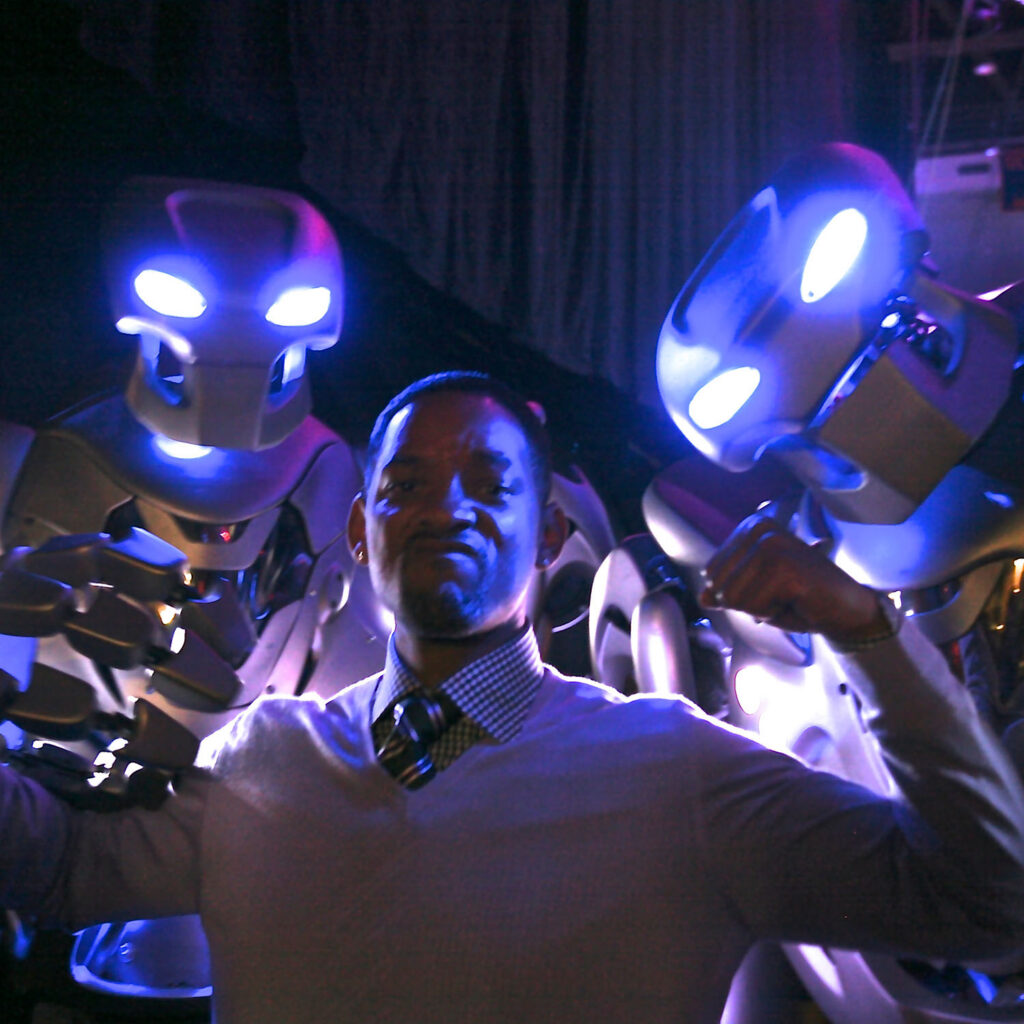 Will Smith stands between two titan the robot's, flexing his arm muscles.