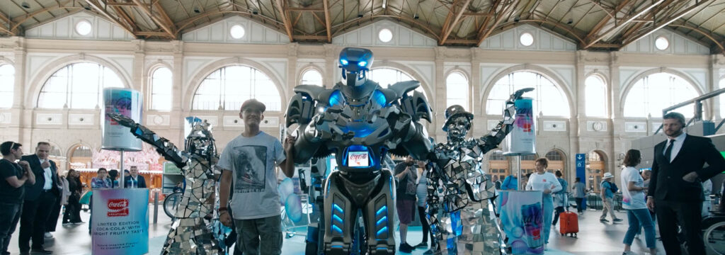Titan the Robot stands with 2 mirror actors at a train station, for the launch of a new coca-cola product -y3000